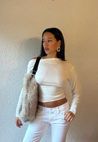 Woman with long dark hair wears white long-sleeve crop top and pants, carrying beige faux fur shoulder bag, looking sideways against textured wall.