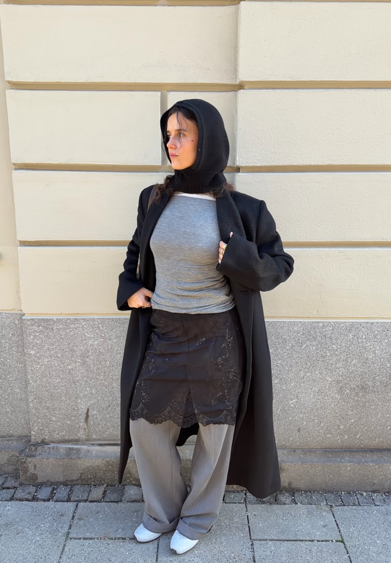 Black long coat with a hood, grey fitted shirt, sheer black skirt with lace hem, loose grey trousers, and white shoes. Wall in background.