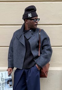 Grijze wollen jas met een hoge kraag, zwarte beanie en zonnebril; houdt een krant vast, draagt marineblauwe broek en een bruine schoudertas.