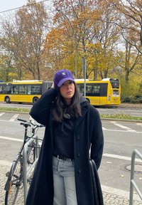 Μωβ μπέιζμπολ καπέλο, μαύρο παλτό, μαύρη φούτερ, ανοιχτό μπλε τζιν, ασημένια ζώνη. Το φόντο περιλαμβάνει δέντρα με φθινοπωρινά φύλλα και ένα κίτρινο λεωφορείο.