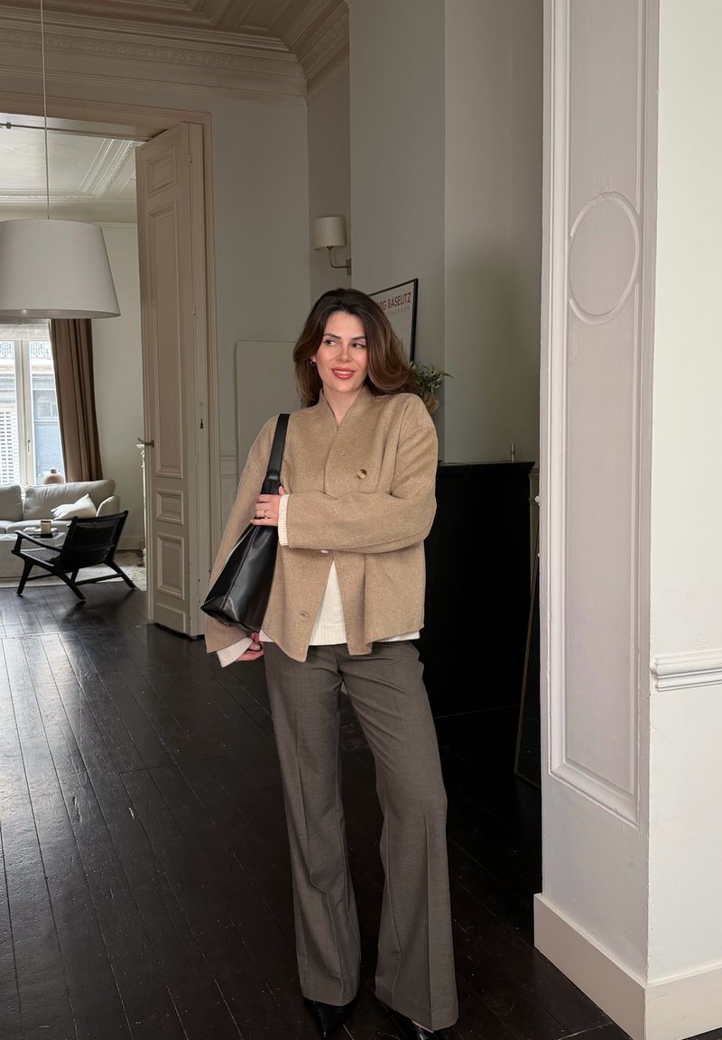 Woman in beige jacket and brown pants holding a black bag, standing in a modern living room with dark wooden floors.