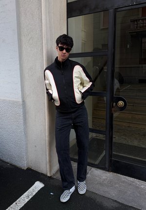 Joven con chaqueta negra y blanca, vaqueros oscuros, zapatos tipo slip-on de cuadros y gafas de sol apoyado en la pared junto a una puerta de cristal.