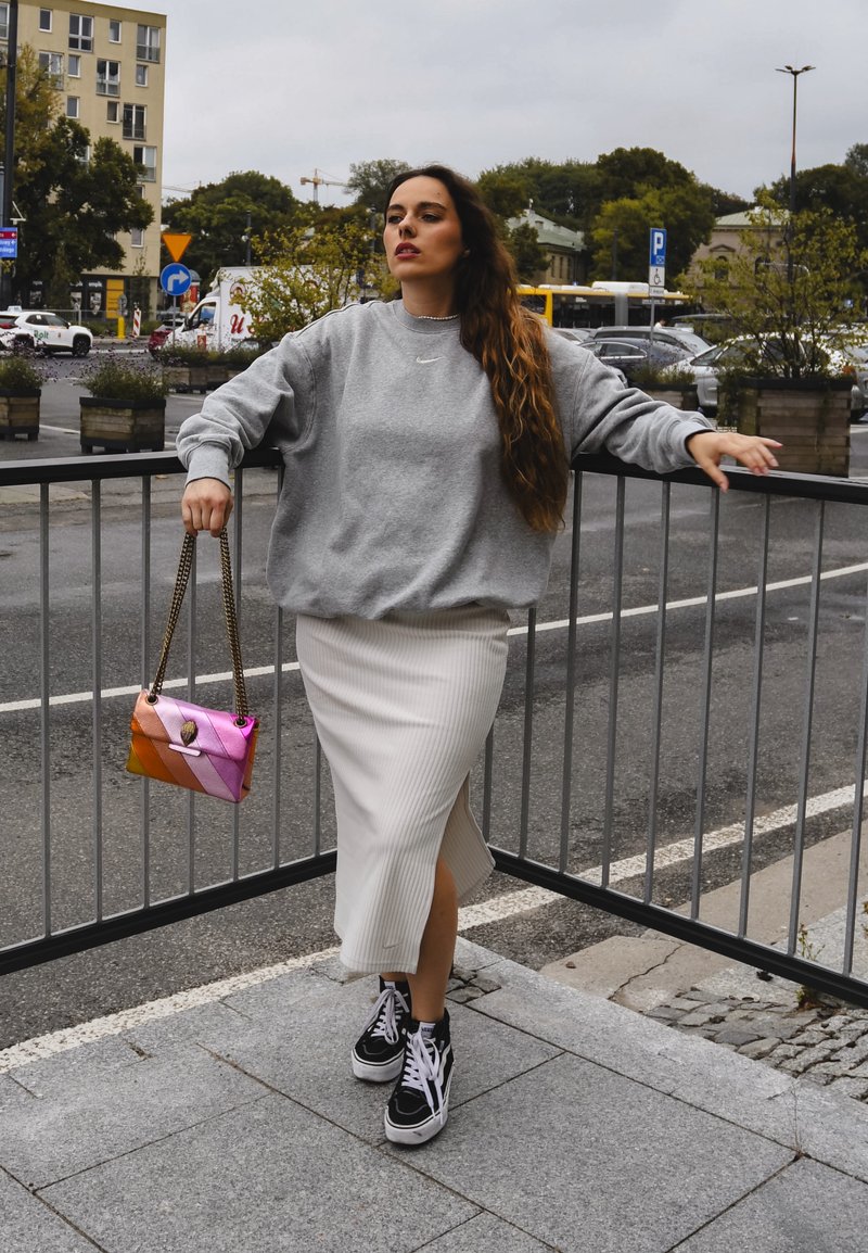Γκρι φαρδύ φούτερ, άσπρη ριμπ μίντι φούστα με πλαϊνό σκίσιμο, μαύρα αθλητικά παπούτσια και μια πολύχρωμη τσάντα ώμου με αλυσιδωτό λουρί.