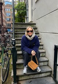 Une personne est assise sur des marches en pierre, portant une veste polaire bleu marine et un pantalon gris, tenant un sac à bandoulière marron, avec des lunettes de soleil et des baskets.