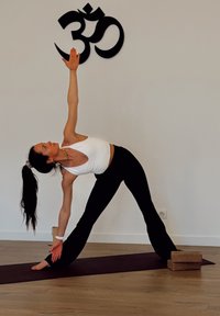 Une personne en crop top blanc et pantalon noir réalise une posture de yoga sur un tapis violet, avec des blocs en liège à proximité, et un symbole Om sur le mur.