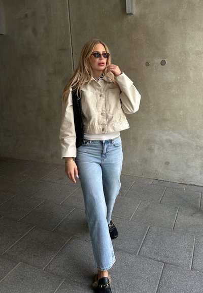 Chaqueta de mezclilla de corte corto en color beige claro sobre una camiseta blanca, combinada con jeans rectos de color azul claro y mocasines negros. Bolsa negra y gafas de sol.