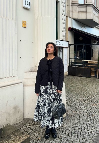 Chaqueta negra acolchada sobre una blusa negra, combinada con una falda larga de estampado blanco y negro. Sostenida por un bolso negro, de pie sobre adoquines.