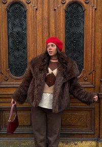 Manteau en fausse fourrure marron, béret rouge, pull à motif losanges beige et marron, couche en tricot crème, pantalon marron, tenant un sac à main bordeaux.