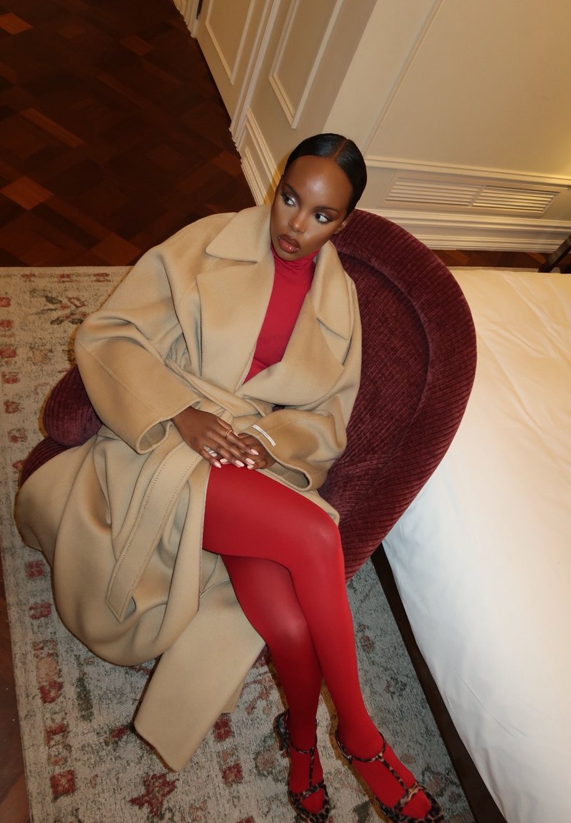 Femme en manteau beige, robe rouge, collants rouges et escarpins imprimé léopard assise sur une chaise bordeaux près d'un lit dans une pièce.