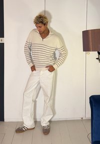 Homme aux cheveux blonds et bouclés portant un pull beige à rayures, un pantalon blanc et des baskets grises, debout à l'intérieur près d'une lampe et d'une chaise bleue.
