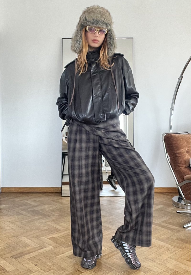 Person iført pels hat, briller, sort læderjakke, brede ternede bukser og sneakers står indendørs på trægulv ved et spejl og en stol.