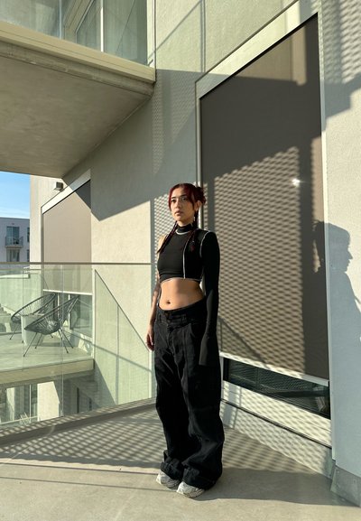 Top corto negro con una manga larga; pantalones negros de talle alto y holgados; zapatillas grises; sombras creadas por el barandal del balcón. Entorno exterior iluminado por el sol.