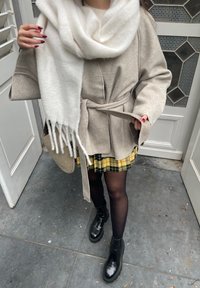 Beige παλτό με ζώνη και φαρδιές μανσέτες, συνδυασμένο με λευκό κασκόλ. Κάτω από αυτό, μια κίτρινη καρό φούστα και μαύρες μποτάκια μέχρι τον αστράγαλο ολοκληρώνουν το ντύσιμο.