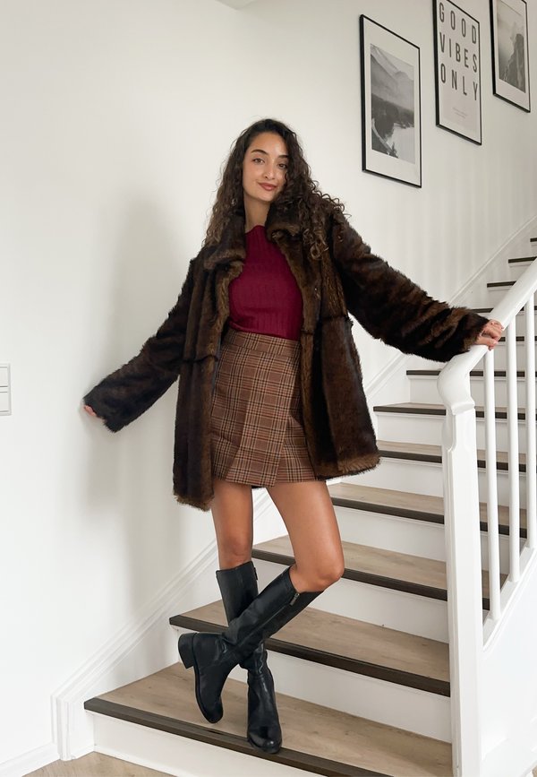 Brauner Mantel aus Kunstpelz, bordeauxroter, gerippter Pullover, karierter Minirock und schwarze kniehohe Stiefel. Lockiges Haar, stehend auf einer Treppe mit weißen Wänden.