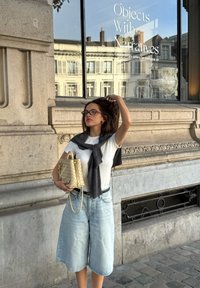 Eine Person in einem weißen T-Shirt, mit einem grauen Pullover über den Schultern und hellblauen Culottes hält eine beigefarbene geflochtene Handtasche vor einem Galerien-Hintergrund.