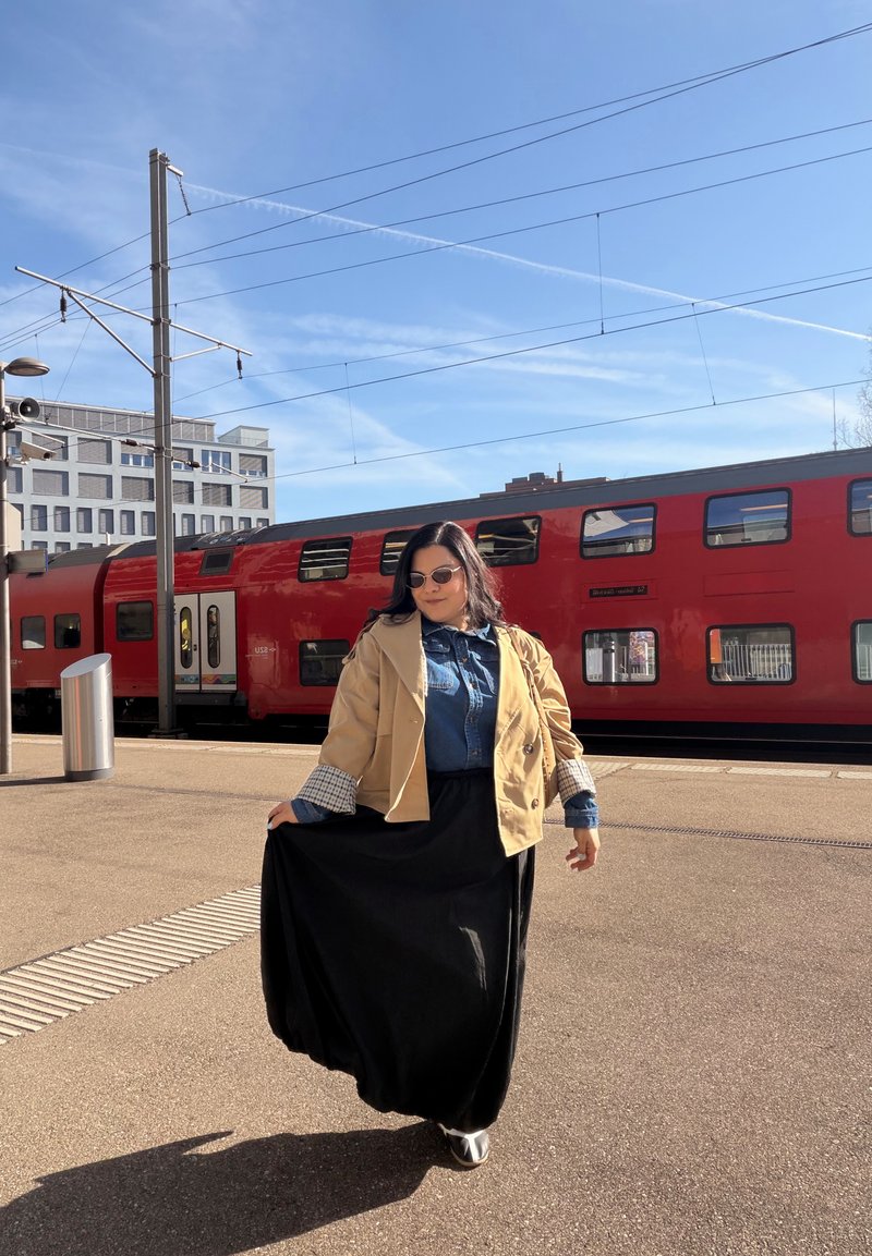 Femme portant des lunettes de soleil, une veste beige, une chemise en denim et une longue jupe noire, debout sur un quai de train avec un train à impériale rouge derrière elle.