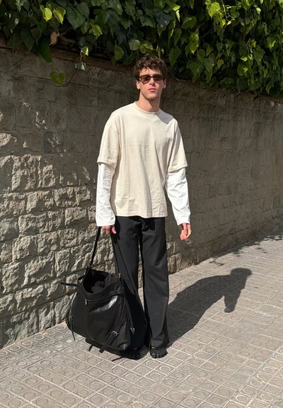Joven vestido con gafas de sol, camisa beige de manga larga con capas, pantalones negros, sosteniendo una gran bolsa negra, de pie en una acera soleada cerca de un muro de piedra.