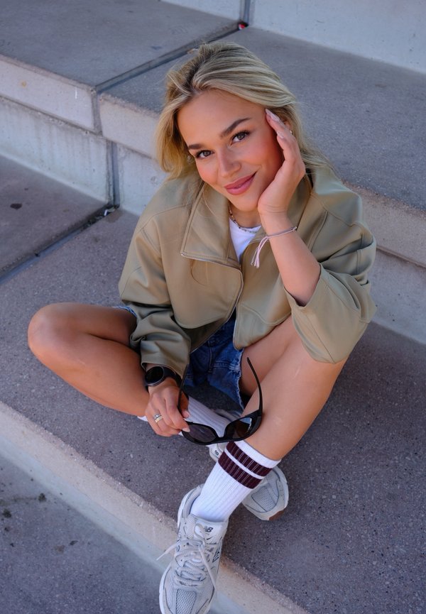 Jeune femme aux cheveux blonds assise sur des marches en béton, souriante et tenant des lunettes de soleil, portant une veste beige, un short en jean, des chaussettes blanches et des baskets.
