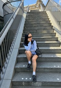 Una persona è seduta su alcuni gradini di pietra, indossando una camicia azzurra chiara con bottoni, pantaloni corti neri, calze bianche e scarpe nere, mentre tiene una borsa nera.