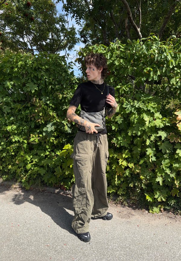 Young person with tattoos wearing a black shirt, khaki cargo pants, and black shoes standing by green leafy bushes in sunlight.