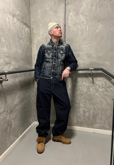 Chaqueta de denim azul con detalles de costura blancos, combinada con jeans oscuros holgados y botas beige. Ambientado contra una pared de concreto gris.