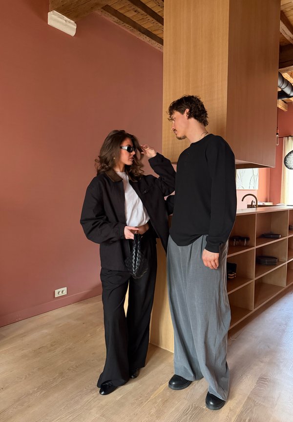 Žena v čiernych slnečných okuliaroch a odeve si upravuje vlasy, zatiaľ čo muž v čiernej košeli a sivých nohaviciach stojí oproti nej v modernej kuchyni.