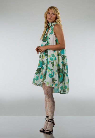 Vestido blanco con estampados florales en azul y verde, diseño acampanado, cuello alto y un lazo en el cuello. Lleva medias con patrón de encaje y tacones negros.