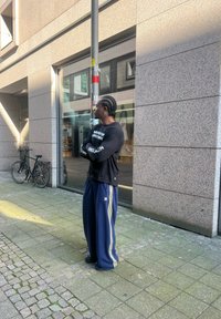 Άνδρας με πλεξίδες φορώντας μαύρη μπλούζα με μακριά μανίκια και μπλε παντελόνι Adidas στέκεται με τα χέρια στα�υρωμένα σε πεζοδρόμιο της πόλης κοντά σε ποδήλατα.