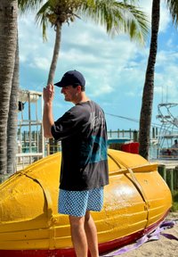 Jeune homme en casquette noire et short à motifs ajustant son chapeau à côté d'un bateau jaune retourné près des palmiers et de la marina par une journée ensoleillée.