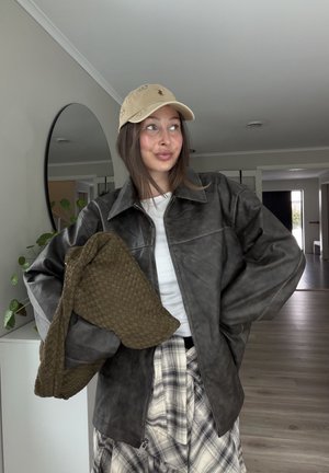 Mujer joven con gorra beige, chaqueta de cuero gris extragrande, falda de cuadros, sosteniendo un gran bolso tejido verde oliva en un espacio interior.