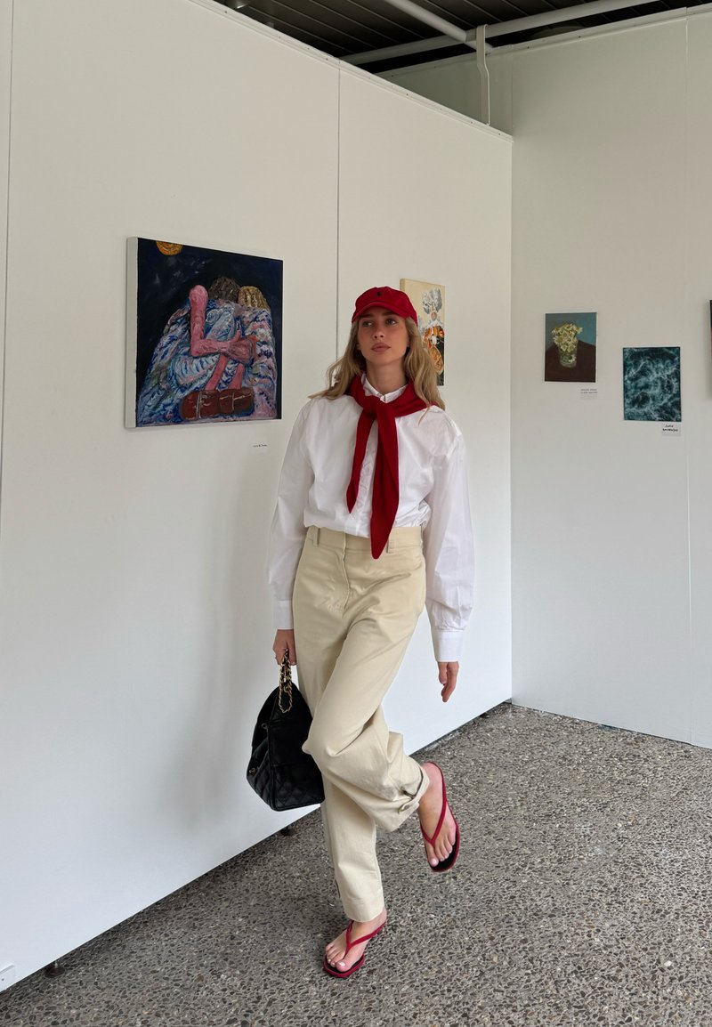 Femme marchant dans une galerie d'art portant une chemise blanche, un pantalon beige, une casquette et une écharpe rouges, des tongs rouges, portant un sac à main noir, avec des tableaux sur des murs blancs.