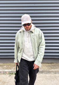 Muž v svetlozelenej bunde, sivom kapucňovom pulóvri, béžovej čiapke, slnečných okuliaroch a čiernych nohaviciach kráča po chodníku vedľa sivej vlnitej kovovej steny, s čiernou taškou na ramene.