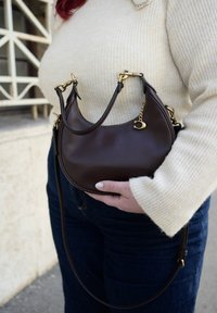 Barna bőrtáska két rövid fogóval és állítható pánttal. Aranyszínű fém díszítéssel és enyhe fényű felülettel. Bordázott krémszínű pulóveres háttér.