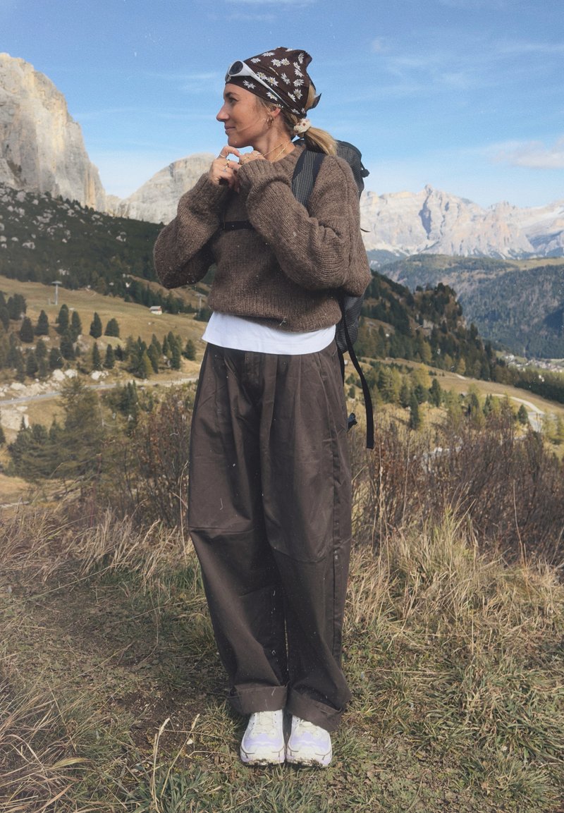 Brown knitted sweater over a white shirt, loose dark brown trousers, and light-colored sneakers. A patterned scarf and a backpack.