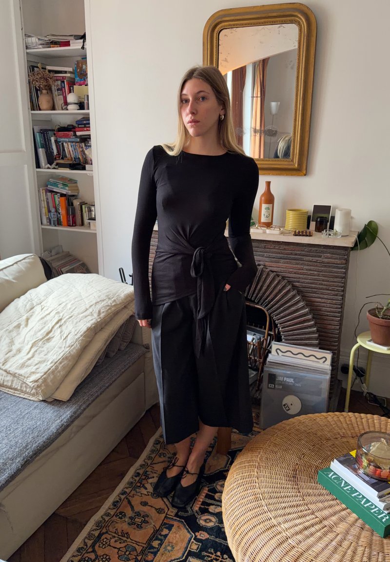 Donna con maglia nera a maniche lunghe e pantaloni ampi in piedi in una stanza con tappeto a motivi, tavolo in vimini, divano, libreria e camino con specchio incorniciato in oro.