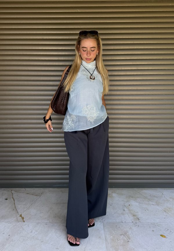 Joven mujer con cabello largo y rubio que lleva una camiseta sin mangas blanca, pantalones negros de pierna ancha, sandalias negras y lleva un bolso marrón.
