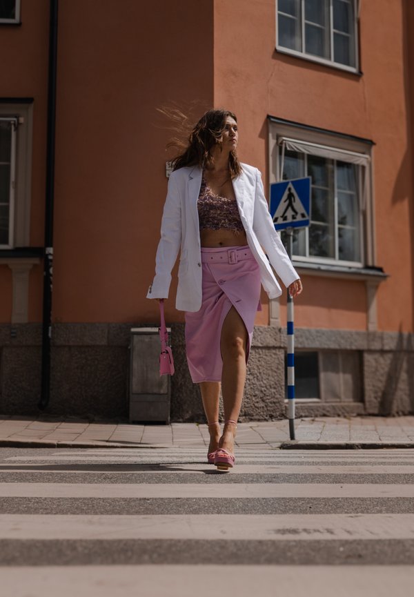 Blazer blanco, top corto morado, falda envolvente rosa con cinturón, bolso rosa y sandalias rosas, caminando por un paso de peatones con un fondo de pared durazno.