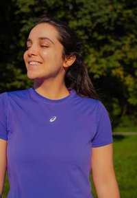 Mlada žena u ljubičastoj sportskoj majici s zatvorenim očima i osmijehom, stoji na otvorenom s zelenim drvećem i travom u pozadini.