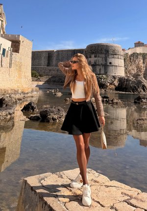 Mujer con cabello largo que lleva gafas de sol, blusa blanca, falda negra y chaqueta beige, de pie sobre una piedra cerca del agua y una fortaleza histórica bajo un cielo despejado.