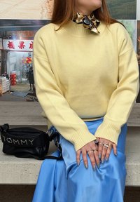 Femme assise portant un pull jaune, une jupe en satin bleu, un foulard à motifs autour du cou, des ongles gris, plusieurs bagues, et un sac à main noir Tommy sur un banc.