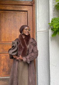 Brun semsket trenchcoat med pelskrage, lagd over en beige kjole. Tilbehør inkluderer et brunt skjerf og briller med klare rammer. Trebakgrunn.