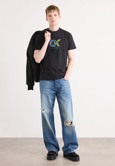 Camiseta de algodón negra con un colorido logo de "OK", combinada con jeans desgastados de un azul claro y zapatos negros gruesos, sosteniendo una chaqueta negra.