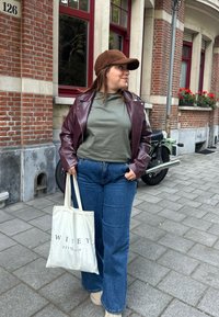 Mørkerød læderjakke, olivengrøn t-shirt, flagrende blå denim jeans, beige sko og en lys tote taske med "WILEY EST. 2024" trykt.