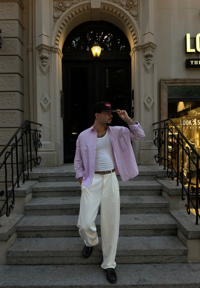 Top blanco de tirantes superpuesto con una camisa a rayas rosa claro, pantalones de pierna ancha crema, zapatos marrón oscuro y una gorra negra con texto rojo, de pie en unas escaleras.