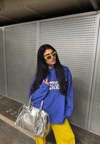 Sweatshirt azul oversized com letras brancas, óculos de sol amarelos brilhantes e uma bolsa prateada texturizada. Calças amarelas completam o visual.