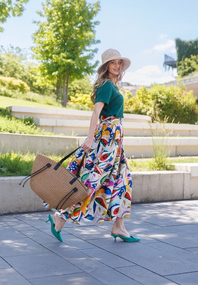 Mujer con sombrero de sol, top verde y falda floral colorida caminando al aire libre llevando un gran bolso tejido bajo un cielo soleado.