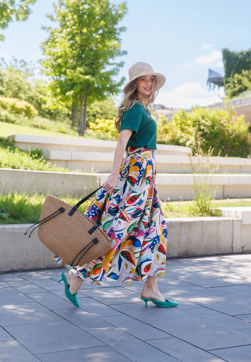 Femme portant un chapeau de soleil, un haut vert et une jupe fleurie colorée, marchant à l'extérieur avec un grand sac à main tressé sous un ciel ensoleillé.