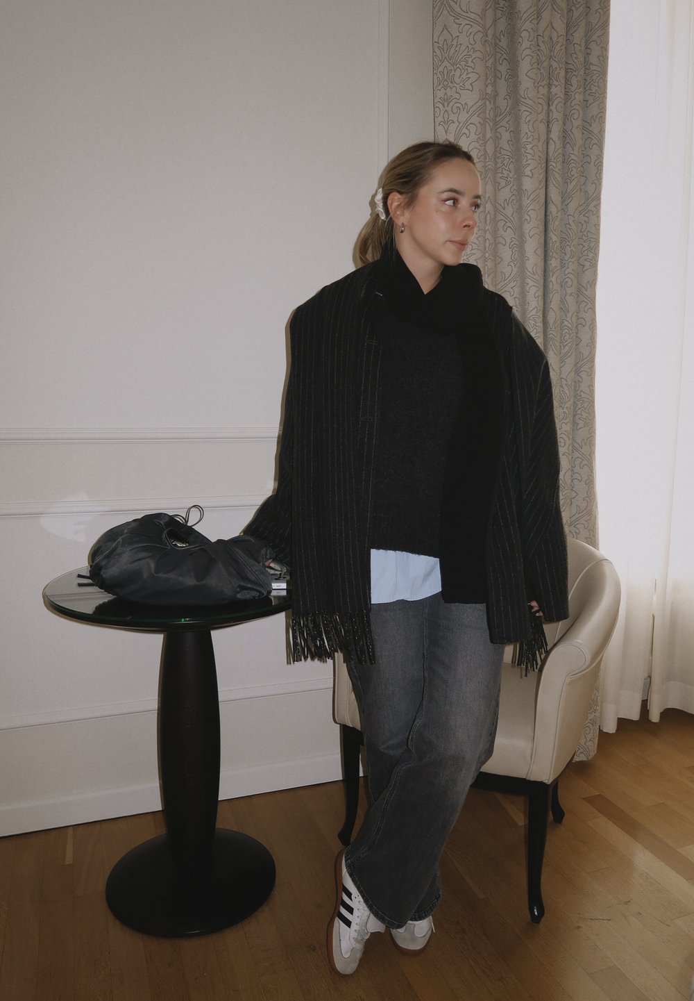 Châle rayé noir et gris avec des franges, pull sombre, chemise bleu clair, jean gris et baskets blanches à rayures noires, reposant sur une table.