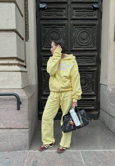 Sudadera de algodón amarilla con logo blanco, pantalones de chándal a juego y zapatillas deportivas marrones. Mochila negra con accesorios y una botella de agua.