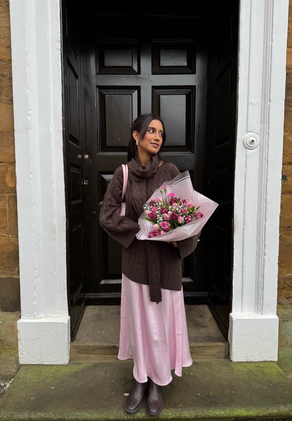 Femme debout dans un cadre de porte tenant un bouquet rose, portant un pull marron, une jupe rose et des bottes, regardant sur le côté.